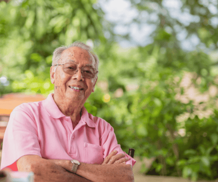 Elderly man in a pink shirt smiles warmly with arms crossed, seated outdoors amidst lush greenery, conveying a feeling of contentment and tranquility.
