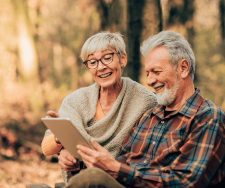 Hs2 pic-min (1) Elderly couple sitting outdoors, smiling while looking at a tablet. The autumn setting with warm tones conveys a joyful and relaxed mood.