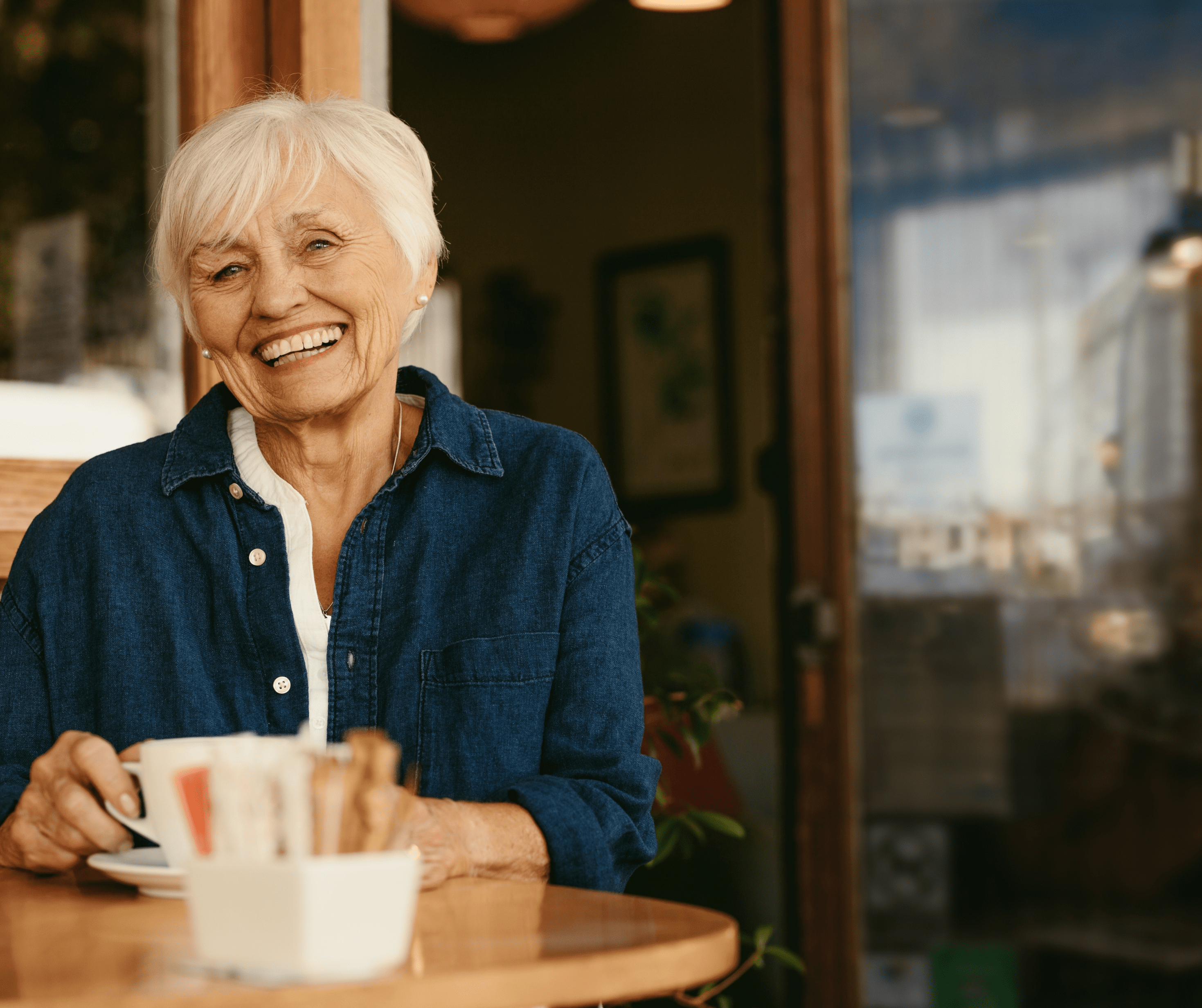 Elderly woman with white hair and a denim shirt smiling warmly while holding a coffee cup at a cozy cafe table, conveying a sense of contentment.