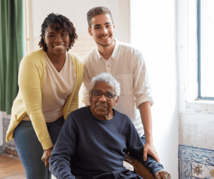A smiling young woman and man stand beside an elderly man seated in a chair, indoors. The atmosphere is warm and familial, with natural light.
