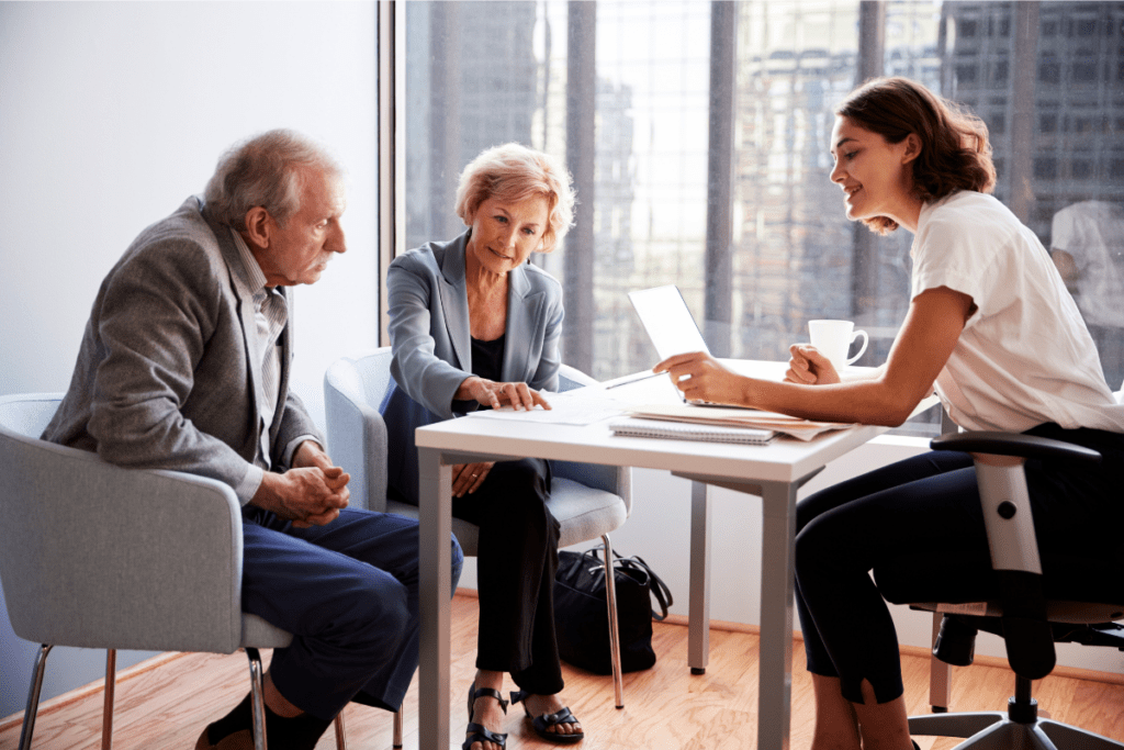 A young professional woman discusses documents with an older couple in a bright office. The mood is collaborative and focused.