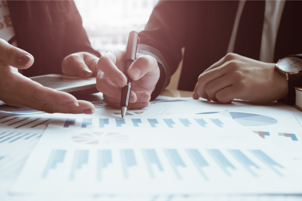 Close-up of hands pointing with a pen at graphs and charts on a table, suggesting a business meeting or discussion. The tone is collaborative and analytical.