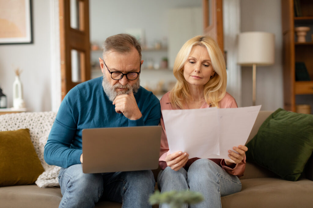 Elderly couple sitting on a sofa, focusing intently. The man uses a laptop while the woman holds papers. The setting is a cozy living room.