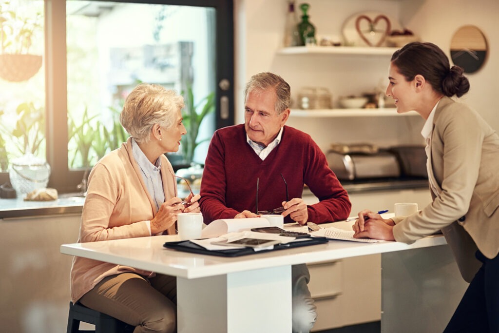 A senior couple and a businesswoman discuss paperwork at a kitchen table. The setting is bright and casual, conveying a friendly, professional tone.