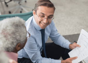 A professional man in a blue shirt and tie smiles, holding papers, as he discusses with two seniors in an office. The mood is friendly and collaborative.