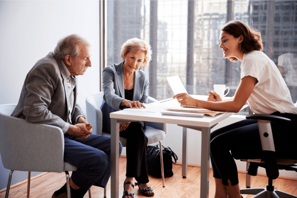 A young professional woman is discussing documents with an older couple in a bright office. They appear focused and engaged in the conversation.