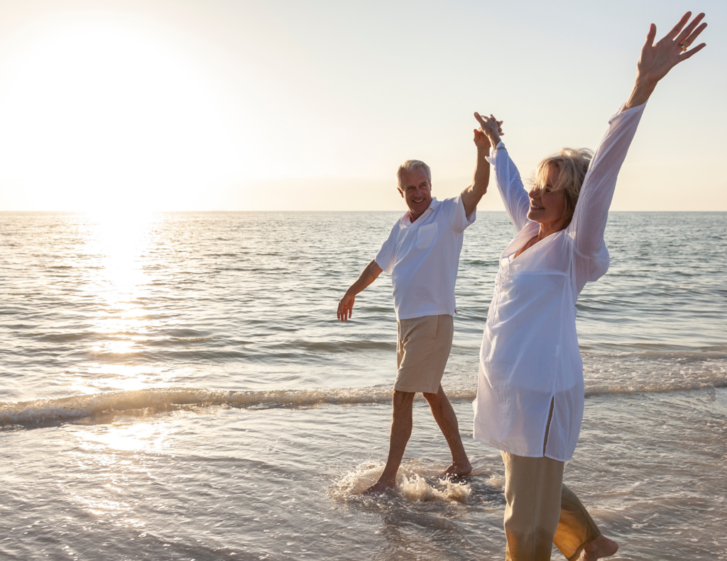 Elderly couple joyfully walking on the beach at sunset, holding hands. The woman raises her arms in delight, the ocean waves gently lapping at their feet.