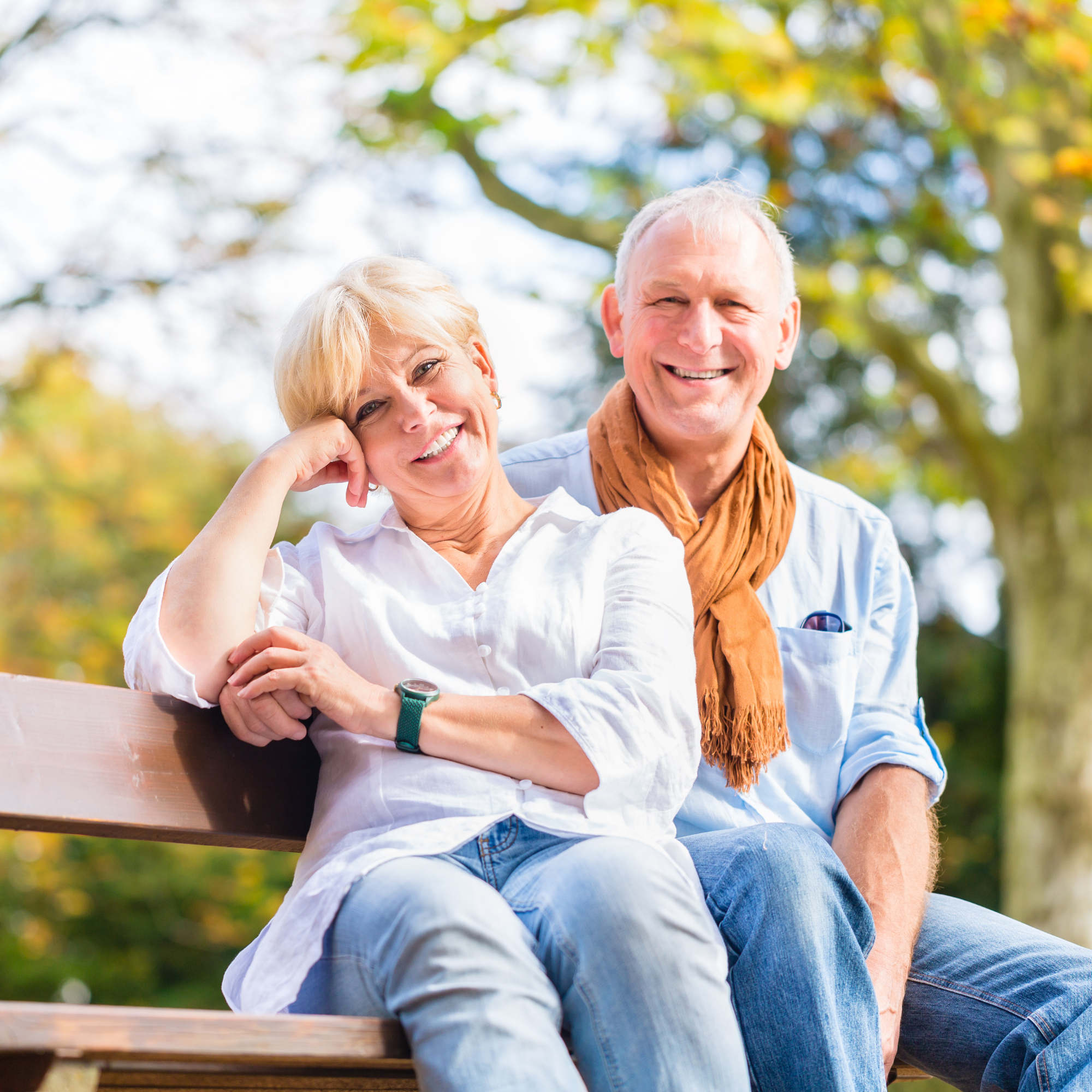 A smiling older woman and man sitting together on a bench with the man arm around the woman's waist. They have relieved and happy expressions and body language since they just got all the reverse mortgage information they needed.